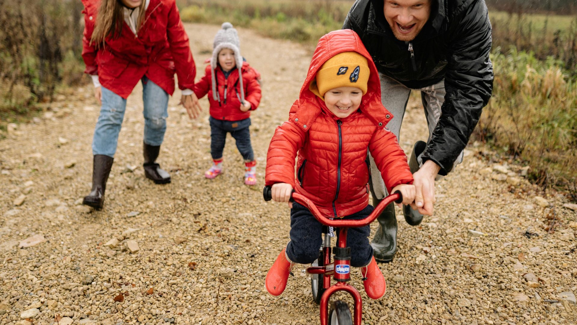 little boy learning to ride his bike while dad holds the bike steady, while mum and young sibling are just behind him cheering