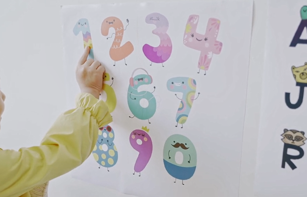 poster of child like illustrated colourful numbers 1-9 pinned up on a wall, with a child wearing yellow pointing to the number 1