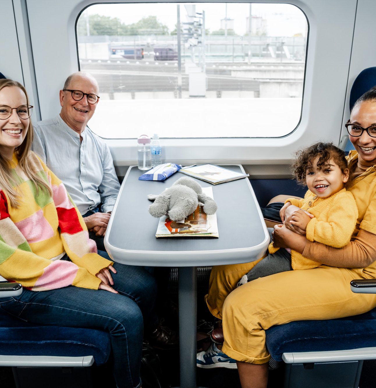3 adults sat around a 4 person seating area on a train, a little girl is sat on her mothers lap, both wearing yellow.